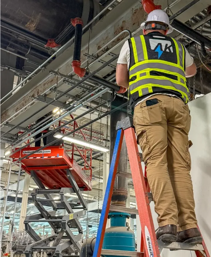 AK Technologies technician in a hi-viz vest installing overhead cable tray from a ladder on a commercial jobsite